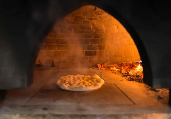 Wnętrze pieca do pizzy z palącym się drewnem i surowym ciastem na kamiennej podłodze. Dym unosi się wokół.