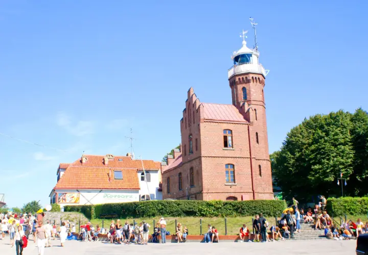 Urokliwa latarnia w Ustce, otoczona zielenią i tłumem turystów.