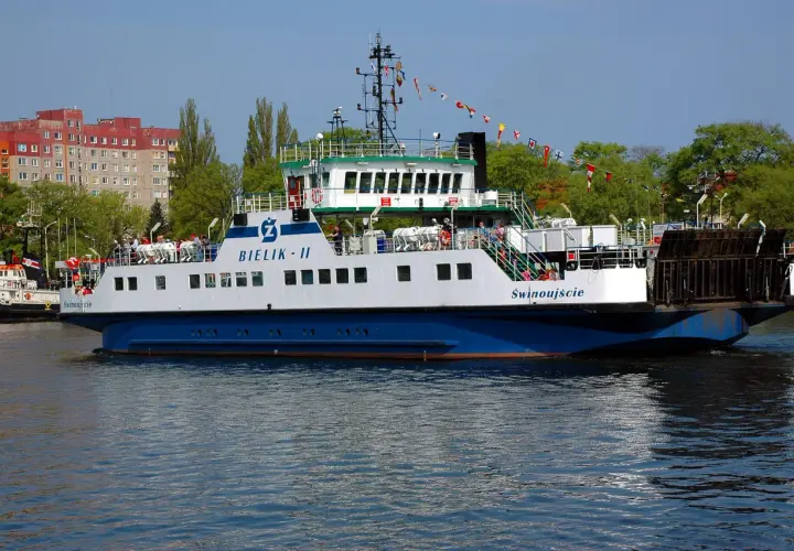 Prom "Bielik II" z pasażerami na pokładzie, płynący po wodzie, z budynkami w tle.