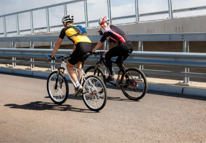Dwóch rowerzystów jedzie obok siebie po moście. Jeden z nich macha do widza.