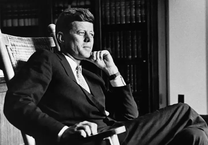 John F. Kennedy sitzt nachdenklich in einem Schaukelstuhl, seine Hand am Kinn. Trotz seiner bekannten **John F. Kennedy Krankheit** strahlt er eine ruhige Entschlossenheit aus.