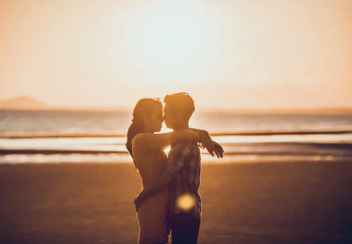 Para zakochanych na plaży o zachodzie słońca. Mężczyzna patrzy kobiecie na usta, obejmując ją.