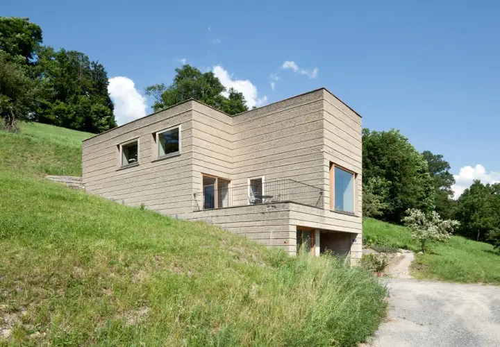 Modernes Haus mit rauer Fassade, das die natürliche Textur von **Lehm als Baustoff** zeigt. Eingebettet in eine grüne Hügellandschaft unter blauem Himmel.