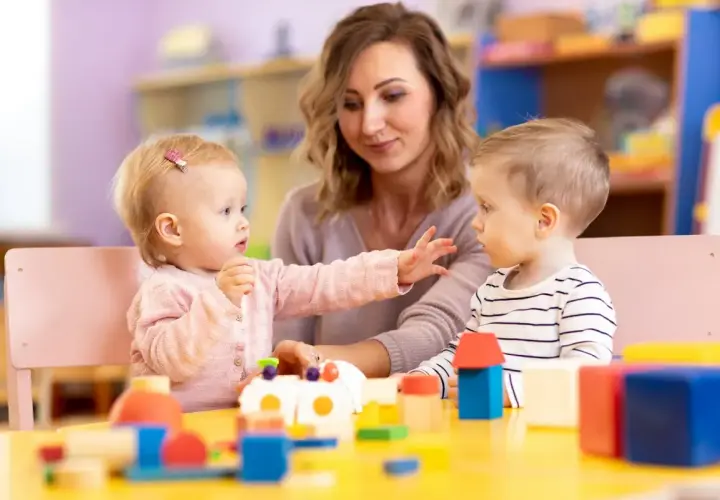 Nauczycielka i dwoje dzieci bawią się klockami przy żółtym stole w żłobku.