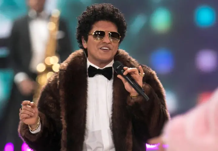 Bruno Mars w futrzanym płaszczu i okularach przeciwsłonecznych śpiewa na koncercie, trzymając mikrofon.