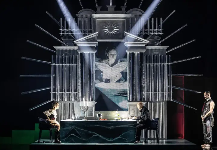 Scena teatralna z projekcją twarzy kobiety, dwójką siedzących przy stole postaci i stojącym mężczyzną. Atmosfera pełna jest teatralnych misteriów.