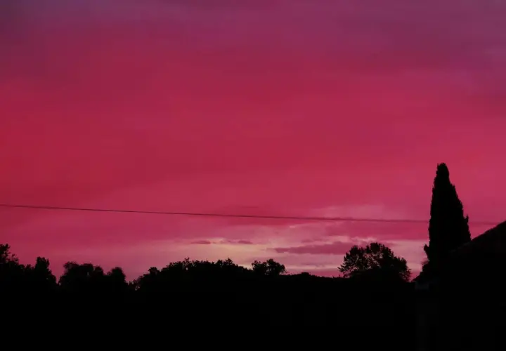Un ciel rouge le soir, teinté de rose, surplombe une silhouette d'arbres et d'une maison.