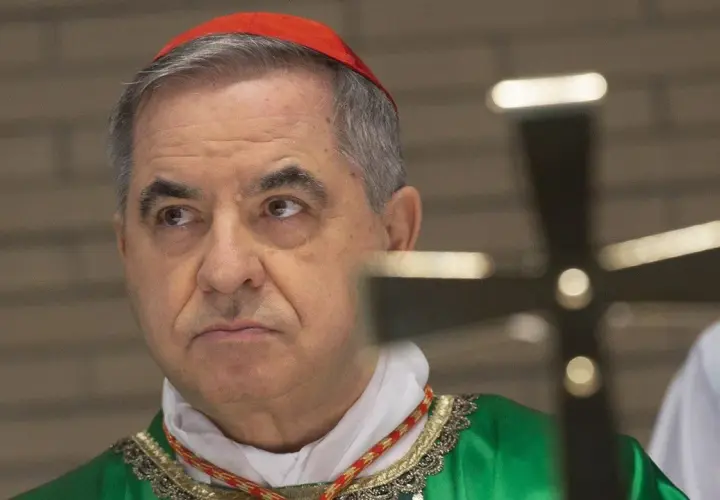 Kardinal Becciu trägt eine rote Pileolus und eine grüne Soutane. Ein Kreuz ist im Vordergrund unscharf.