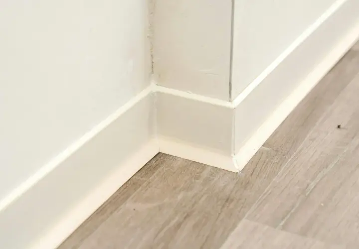 Angolo di un battiscopa in ceramica bianca, pronto per essere incollato su un pavimento in laminato grigio.