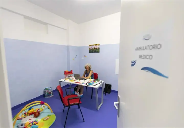 Un ambulatorio medico con una donna al computer, giocattoli e un tappeto gioco. La differenza tra ambulatorio e poliambulatorio è la specializzazione.