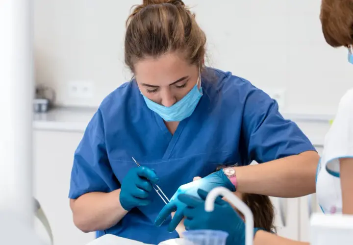 Dentystka w niebieskim fartuchu i masce zakłada opatrunek na zęba pacjentowi.