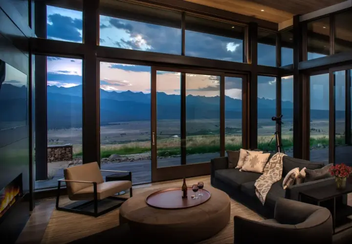Cozy living room in a modern mountain house, featuring a fireplace, plush seating, and expansive windows with a breathtaking mountain vista at dusk.