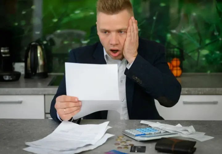 Zaskoczony mężczyzna z papierami i kalkulatorem na stole zastanawia się, jak spłacić debet.