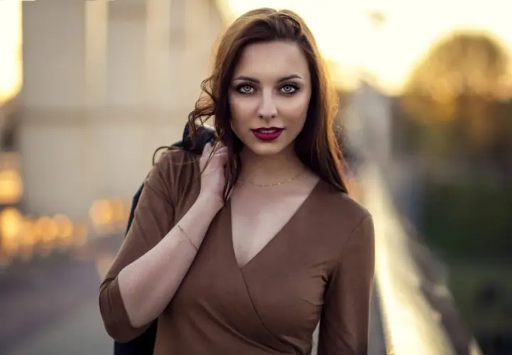 Portret młodej kobiety z burzą brązowych włosów, w brązowej sukience, z kurtką na ramieniu. Ta fotografia ludzi uchwyciła jej spojrzenie.