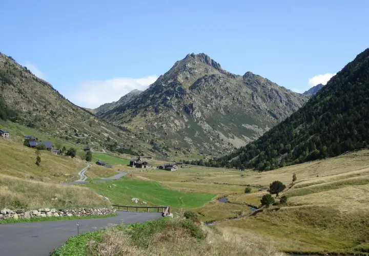 Un camino serpentea hacia el estany de Juclar, un valle andorrano con casas dispersas y un pico imponente.