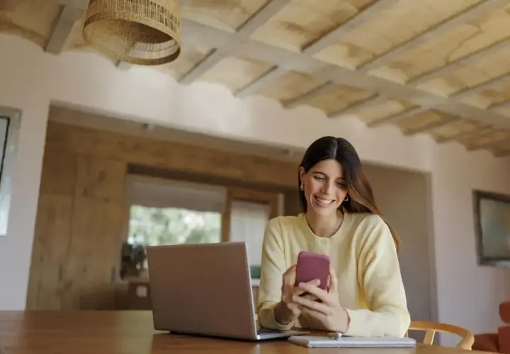 Uśmiechnięta kobieta korzysta z telefonu, sprawdzając mbank księgowanie przelewów. Obok leży laptop.