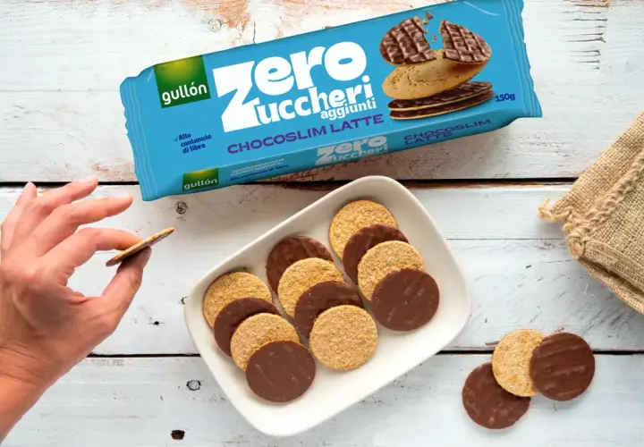 Biscotti senza zucchero Gullón Chocoslim Latte, alcuni ricoperti di cioccolato, su un piatto bianco. Una mano ne prende uno.