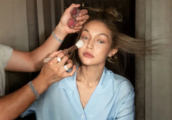 En makeupartist applicerar rouge på en kvinna som håller två små burkar med rouge. Kvinnan har sitt hår uppsatt och bär en ljusblå skjorta.