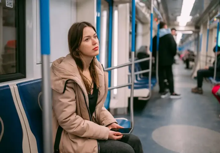 Młoda kobieta w beżowej kurtce siedzi w metrze, trzymając telefon. Jej zamyślona mina sugeruje **wypalenie zawodowe**, być może szuka ulgi lub sposobu na L4.