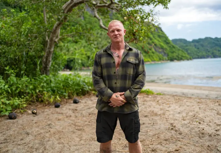 Alexander Strandberg Robinson står på en strand med gröna kullar och vatten i bakgrunden.