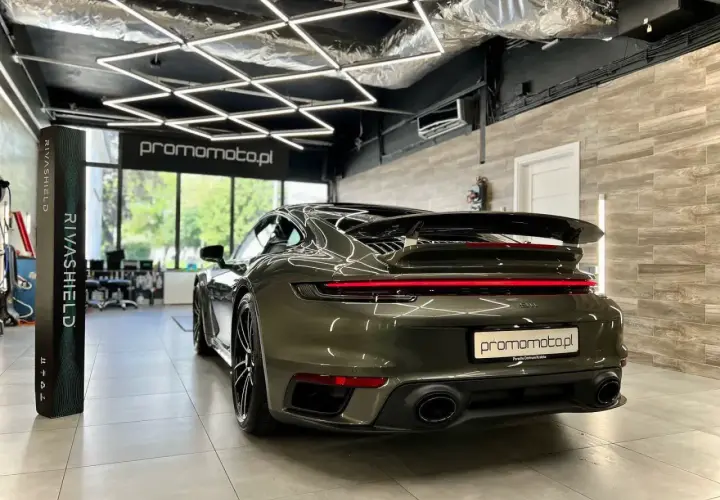 Ciemnozielone Porsche 911 w garażu z nowoczesnym oświetleniem i stojącym obok pionowym banerem.