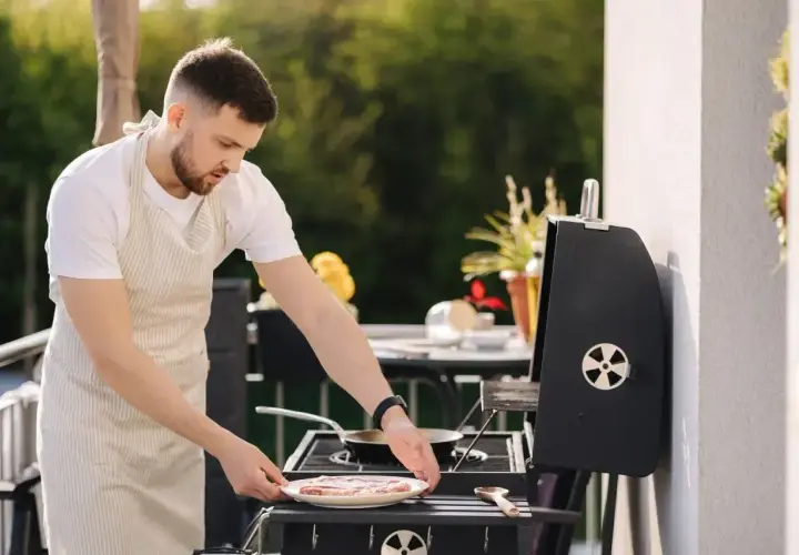 Mężczyzna w fartuchu przygotowuje mięso na grillu na balkonie.