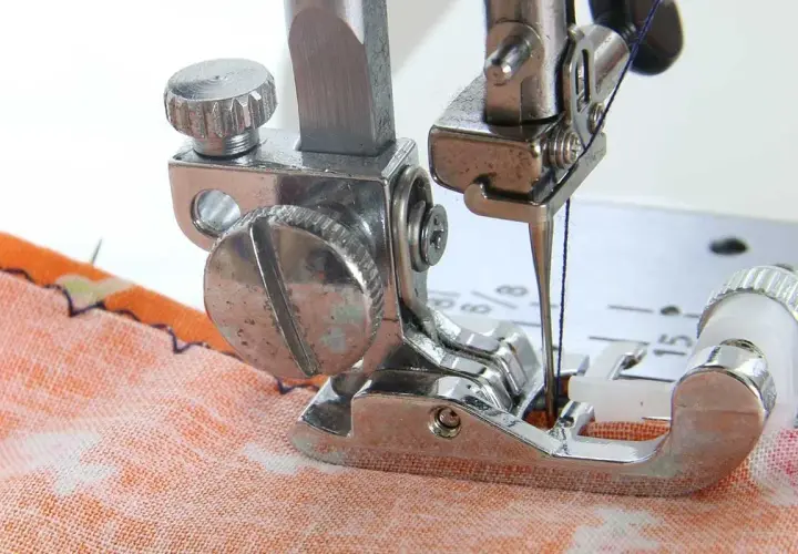 Close-up of a sewing machine needle and blind hem foot working on orange fabric. Learn how to use blind hem foot for invisible hems.