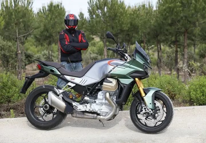Motociclista con casco posa junto a una moto Guzzi Mandello verde y plateada, lista para la aventura en un camino rural.