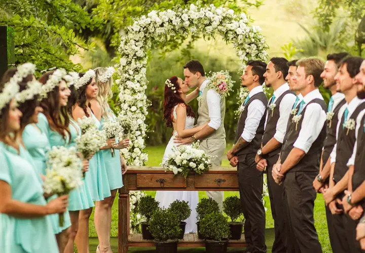 Novios besándose bajo un arco de flores, rodeados de damas de honor y padrinos.