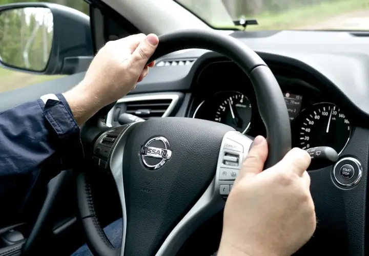 Manos al volante de un Nissan, sintiendo la vibración que indica porque vibra el volante del coche al conducir por un camino rural.