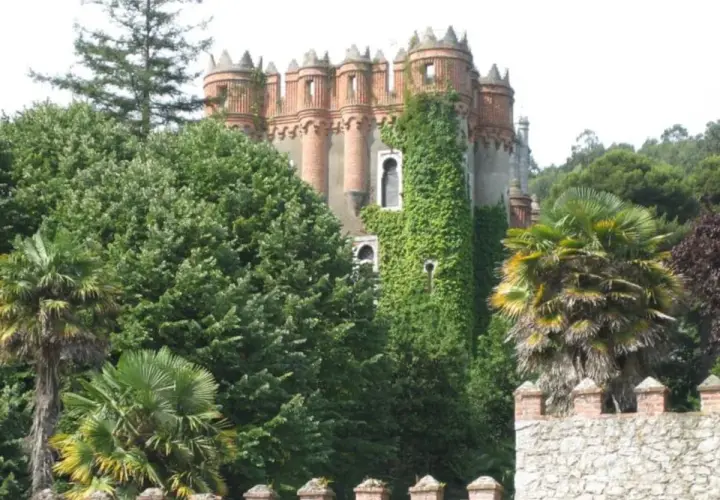 El **palacio de Ocharan**, cubierto de hiedra, se alza entre exuberante vegetación y palmeras, con un muro de piedra en primer plano.
