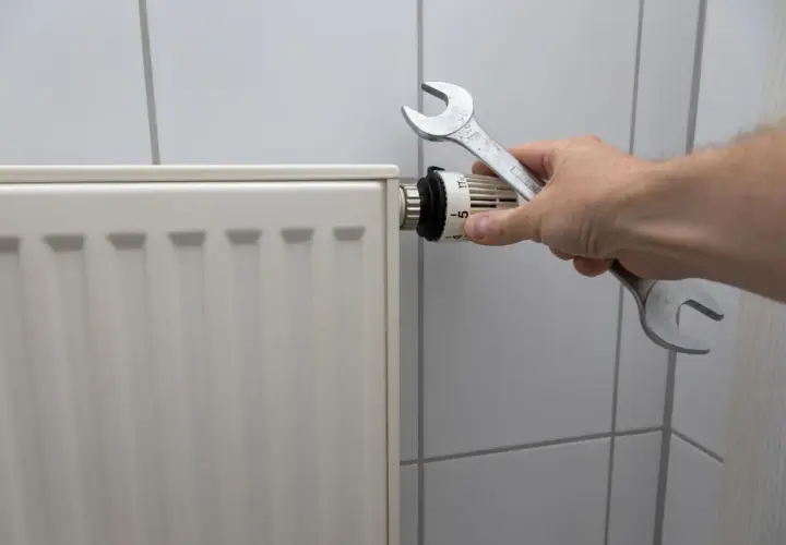 Dłoń z kluczem nastawia termostat, pokazując, jak spuścić wodę z kaloryfera.