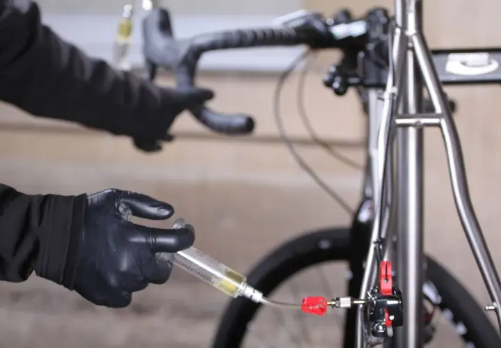 Hands in black gloves perform a bike brake bleed, following the correct brake bleed order to ensure optimal performance.