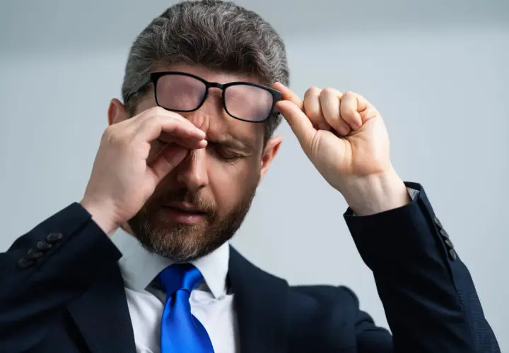 Mężczyzna w garniturze i okularach pociera oko, jakby coś wpadło do oka pod górną powiekę.