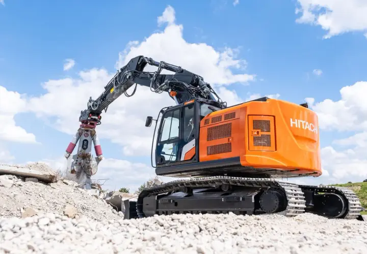 Pomarańczowa koparka do rozbiórek Hitachi z chwytakiem pracuje na gruzowisku pod błękitnym niebem z chmurami.