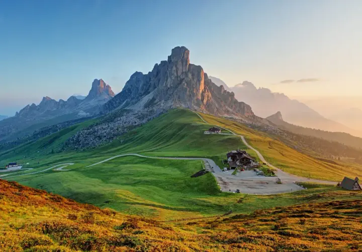A winding road leads to a mountain lodge, perfect for hiking the Dolomites, Italy. Lush green hills meet jagged peaks under a soft sunrise.