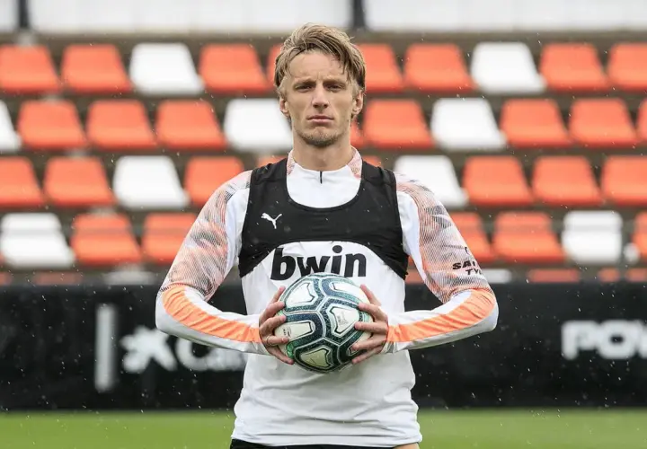 Piłkarz z blond włosami trzyma piłkę, ubrany w białą koszulkę z pomarańczowymi rękawami i czarną kamizelkę z logo bwin. W tle pomarańczowo-białe trybuny.