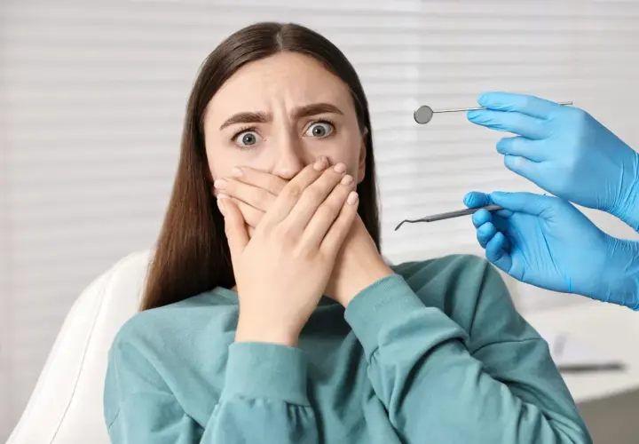 Przerażona pacjentka z dentofobią zakrywa usta. W pobliżu widać ręce dentysty w niebieskich rękawiczkach z narzędziami.