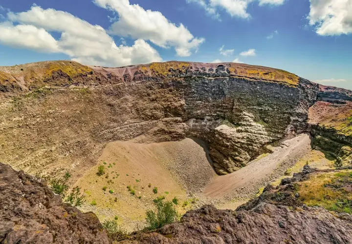 Il cratere del Vesuvio, con le sue pareti stratificate e il cono di detriti al centro, sotto un cielo azzurro con nuvole bianche.