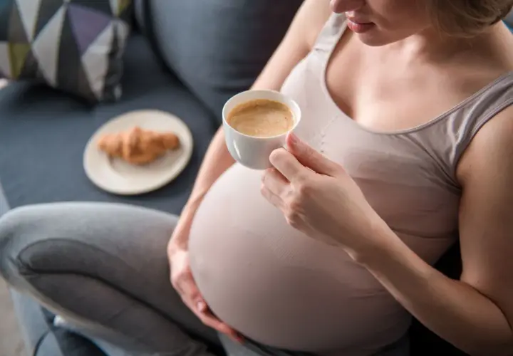 Kobieta w ciąży delektuje się kawą rozpuszczalną, siedząc na kanapie z rogalikiem na talerzyku obok.