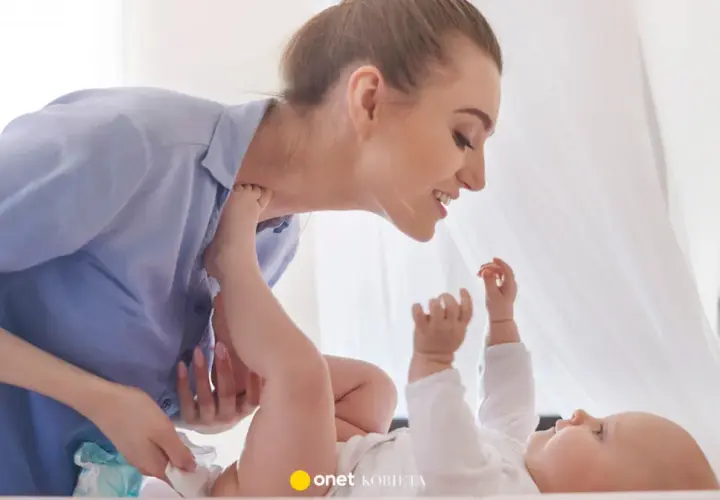 Uśmiechnięta mama pochyla się nad dzieckiem, które leży na plecach. Wykonuje szerokie pieluchowanie, delikatnie unosząc nóżki malucha.