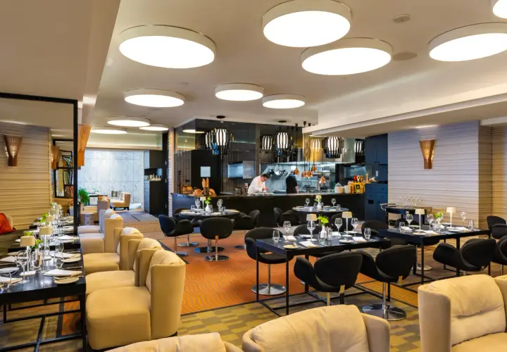 Elegancka brasserie z widokiem na kuchnię, gdzie szef kuchni przygotowuje dania. Stoliki nakryte do kolacji, wygodne fotele i nowoczesne oświetlenie tworzą przytulną atmosferę.