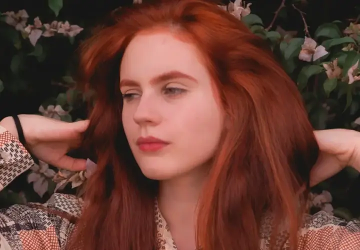 Chica con vibrante pelo pelirrojo, rodeada de flores y follaje, con las manos jugando con su cabello.