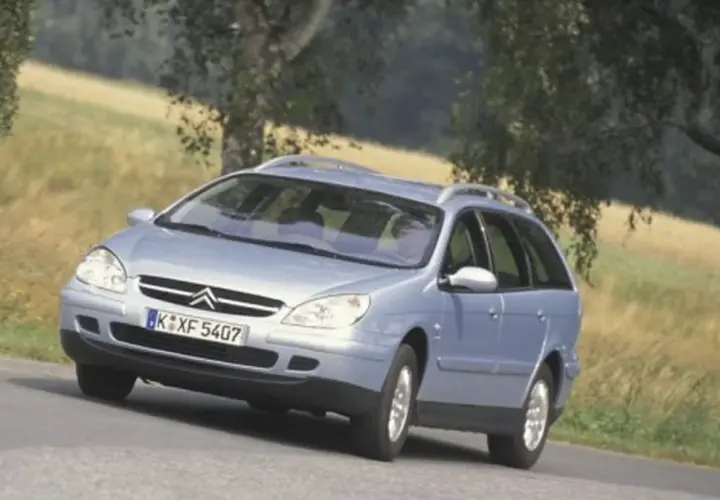 Jasnoniebieskie kombi Citroen na zakręcie drogi, z drzewami i polem w tle.