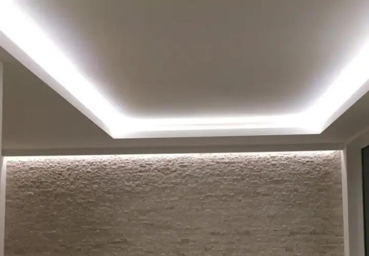 Illuminazione indiretta su velette in cartongesso, che creano un effetto soffuso su una parete in pietra chiara.