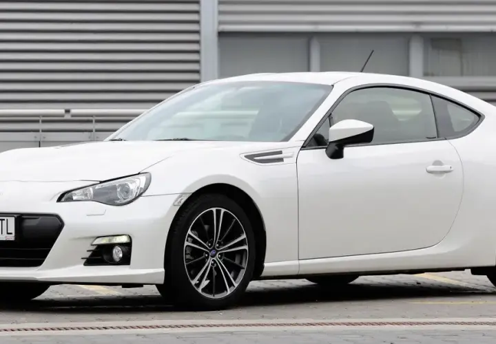 Białe Toyota BRZ z czarnymi felgami stoi przed budynkiem z poziomymi żaluzjami.