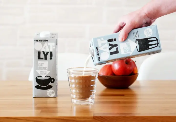 Ręka wylewa mleko dla baristów Oatly do szklanki. Obok stoi karton tego samego napoju.