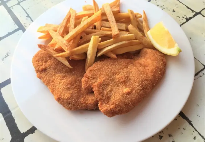 Due cotolette di pollo dorate e croccanti, accompagnate da patatine fritte e uno spicchio di limone su un piatto bianco.