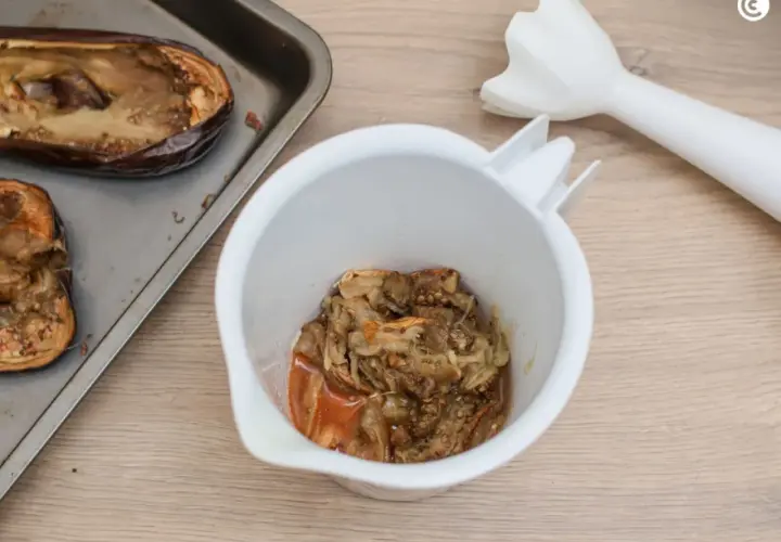 Berenjenas asadas en una bandeja, listas para convertirse en un delicioso hummus de berenjena. Un batidor de mano espera para mezclar.