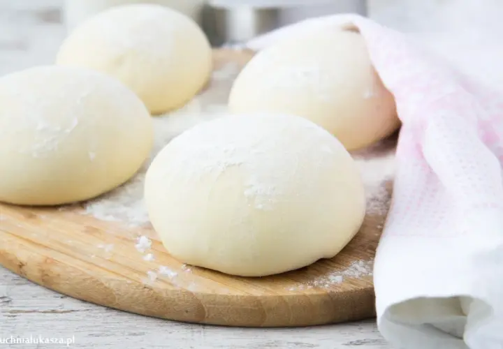 Kulki ciasta, idealne ciasto na pizzę, posypane mąką, leżą na desce. Obok różowy ręcznik.